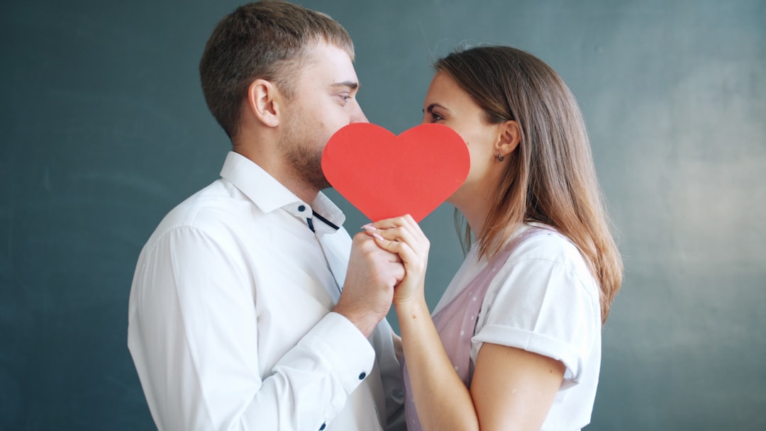 mission-img Beautiful young couple man and woman are kissing hiding behind Valentines card on gray background. Human feelings, millennials and celebrations concept.